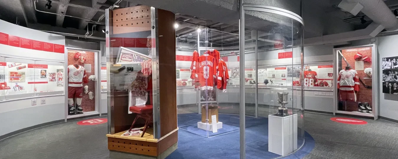 Hockey museum exhibit with red jerseys displayed in glass cases, trophies, and historical photos on walls.