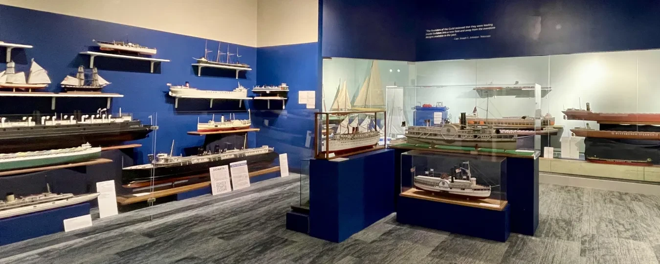 Museum exhibit room with model ships displayed on walls and in glass cases, under dim lighting.