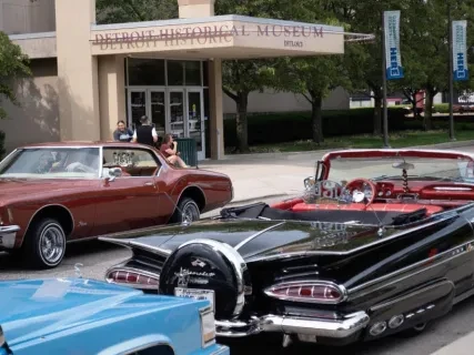 Classic cars parked outside a museum entrance, surrounded by trees and people.
