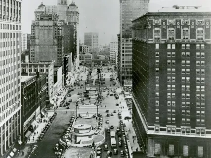 Historic downtown street view with tall buildings, bustling traffic, and vintage cars in black and white photograph.