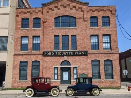Historic brick building with "Ford Piquette Plant" sign, flanked by two vintage cars, one red and one blue, under a clear sky.