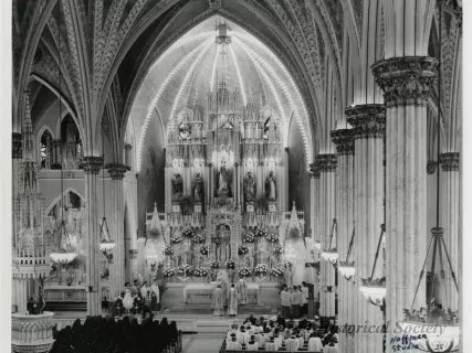 Gothic cathedral interior with vaulted ceilings, ornate altar, and a congregation attending a service. Black and white photograph.