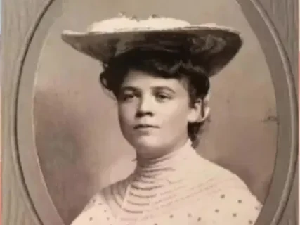 Sepia portrait of a woman wearing a wide-brimmed hat and a high-necked, polka dot dress.