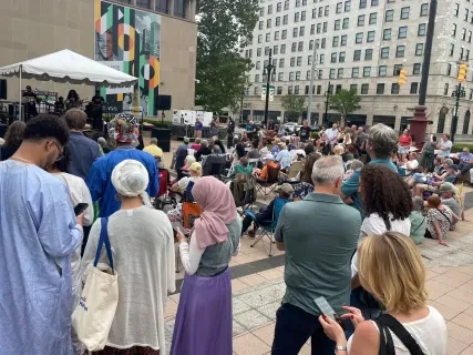 Crowd gathered at an outdoor event in a city square, people sitting and standing, some looking at phones.