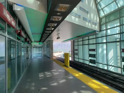 Modern train station platform with glass walls and ceiling, sunlight streaming in, green and yellow signage visible.