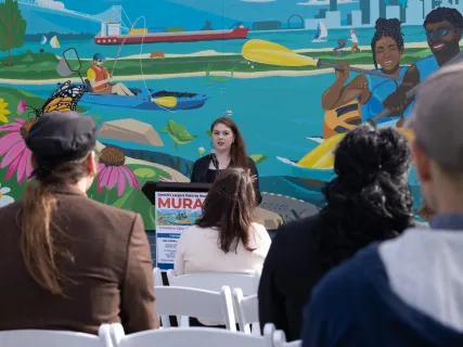 A woman speaks at an outdoor event, with a colorful mural depicting kayakers and nature in the background.