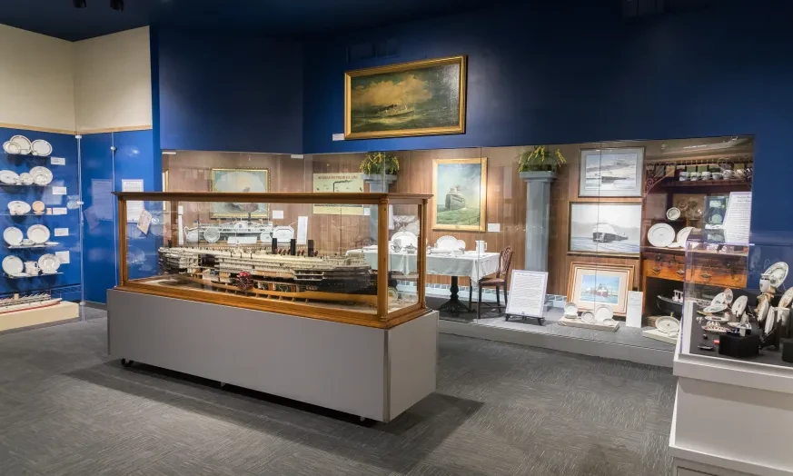 Museum exhibit with a ship model in a glass case, surrounded by maritime paintings and artifacts on the wall.