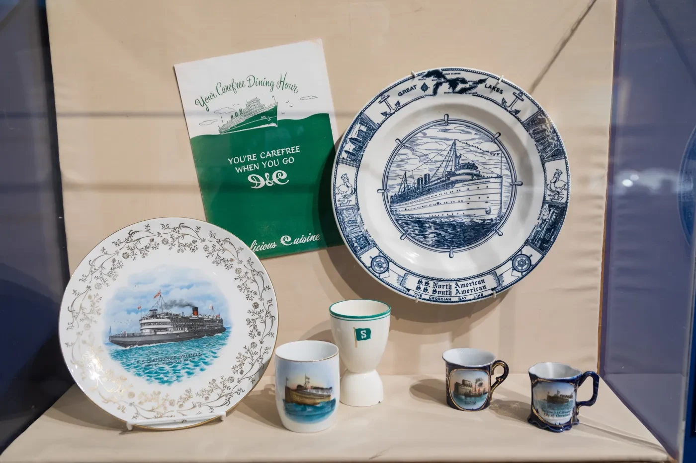 Ceramic plates and mugs with ship illustrations displayed on a shelf.