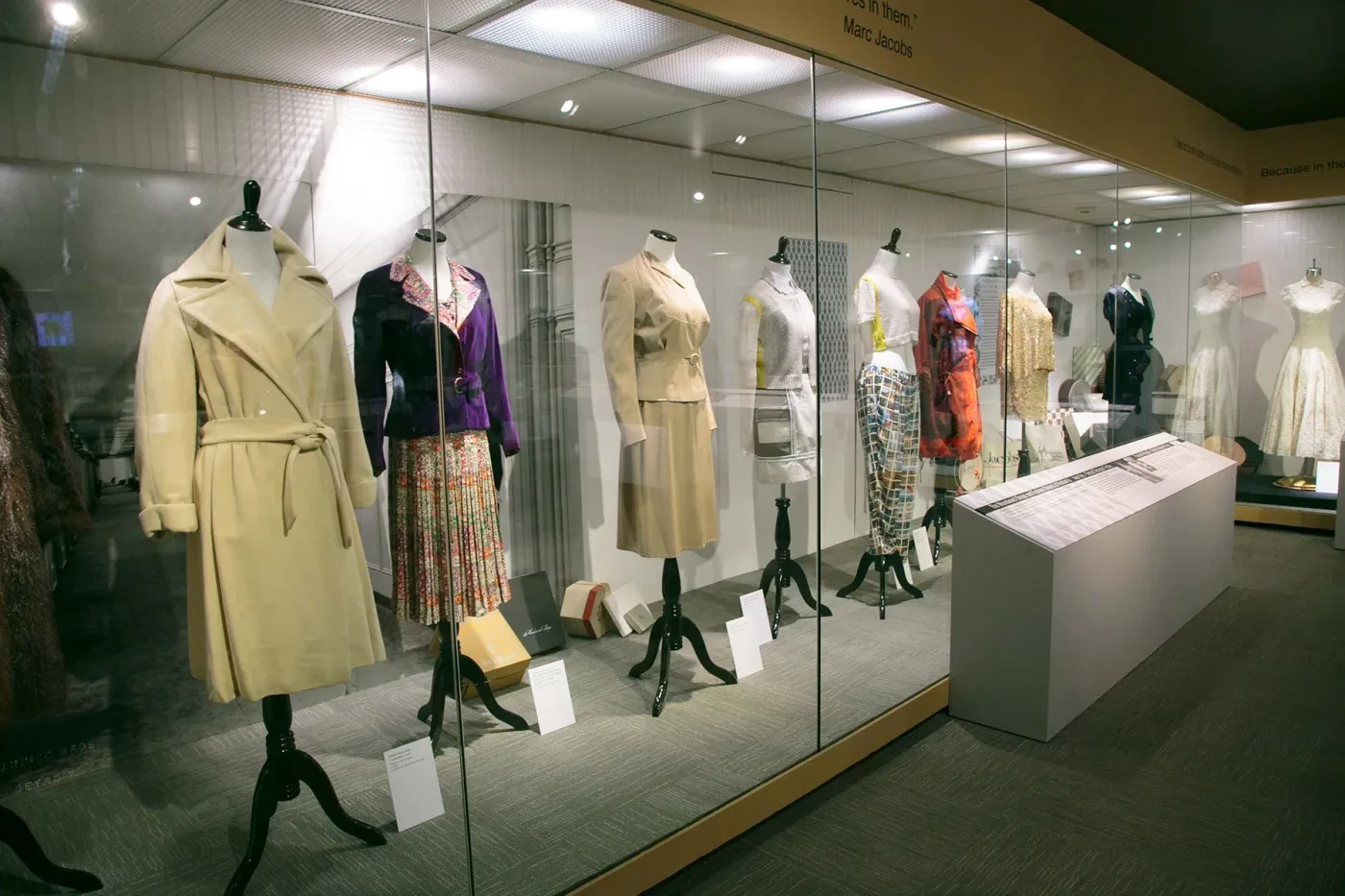 Fashion exhibit with various women's outfits on mannequins in a glass display case.