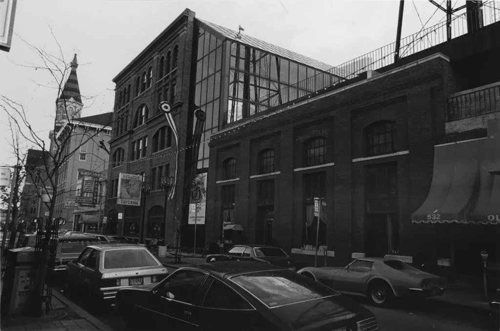 Black and white photo of a city street, lined with parked cars and brick buildings, under a cloudy sky.