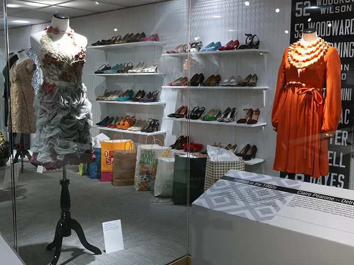 Mannequins with dresses in a shop window, surrounded by shelves of colorful shoes and shopping bags.