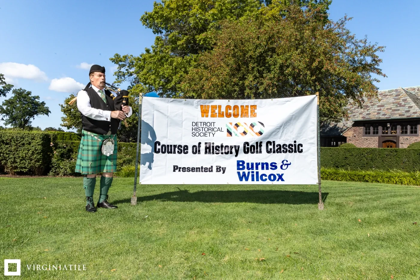 Bagpiper in a kilt stands beside a "Course of History Golf Classic" banner on a sunny day with trees in the background.