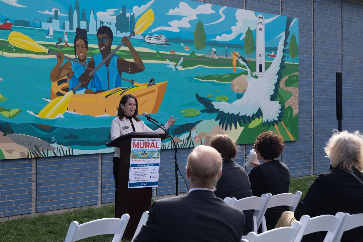 Speaker at a podium with an audience, in front of a colorful mural of people kayaking and nature scenes.