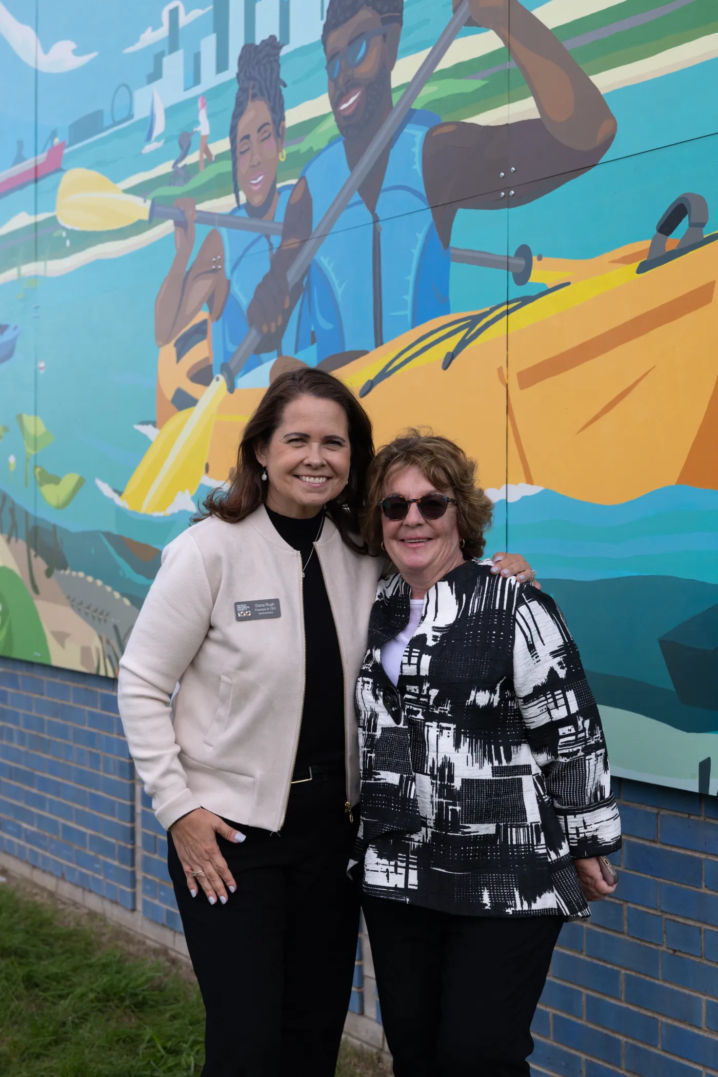 Two women smiling and posing in front of a colorful mural depicting people kayaking.