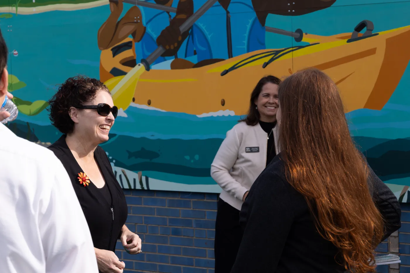 Three people standing and smiling in front of a colorful mural of a boat.