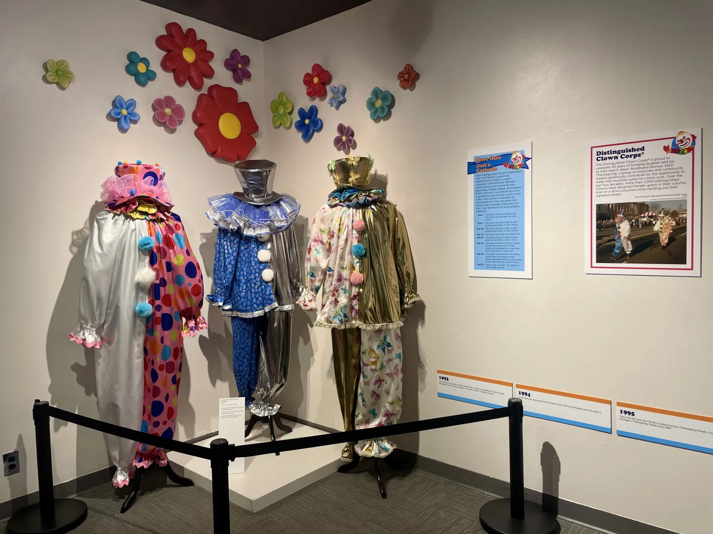 Three clown costumes on display with colorful flower decorations on the wall behind them.