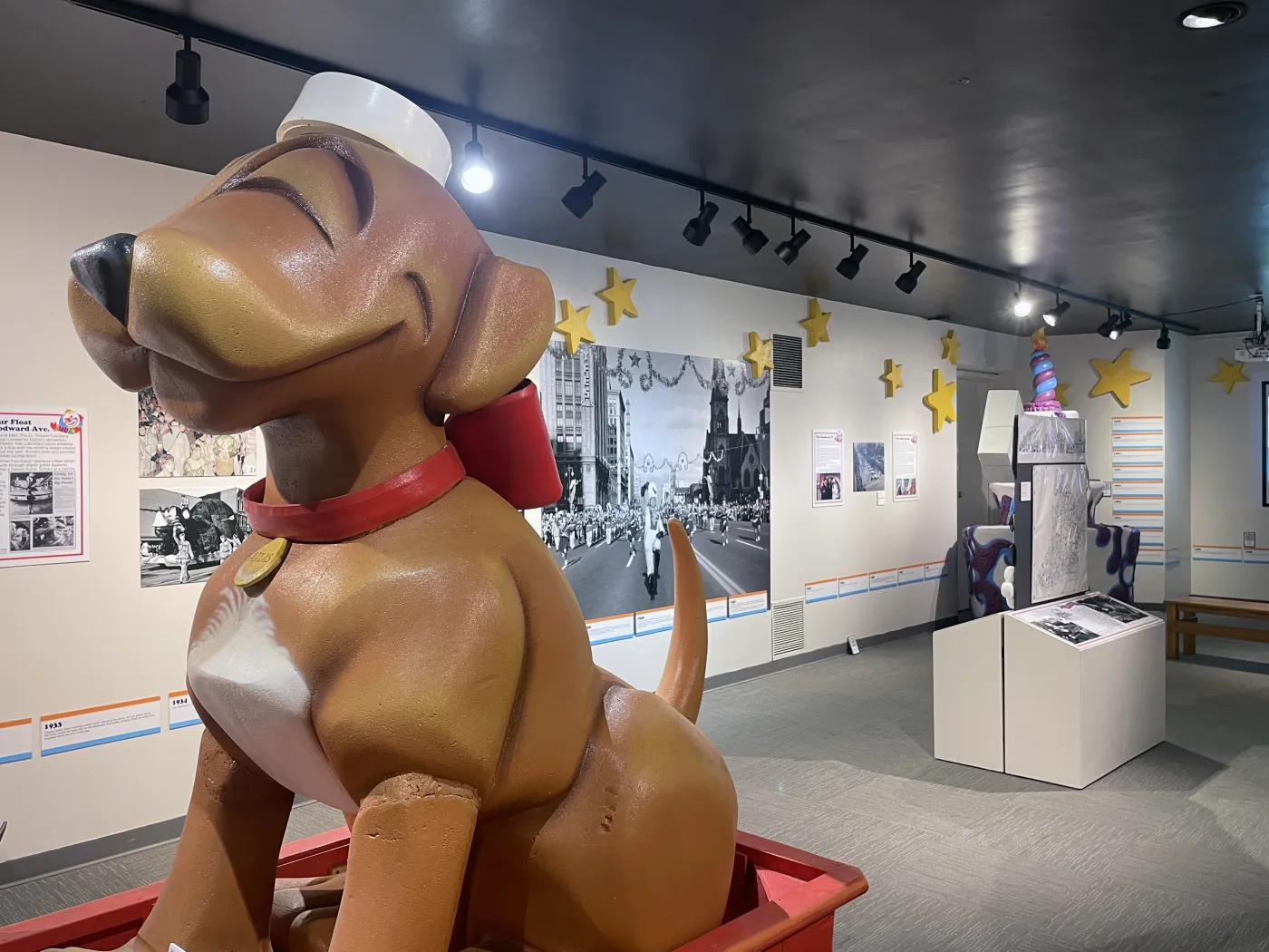 Large smiling dog statue in a sailor hat and red collar inside a museum exhibit with photos and star decorations.