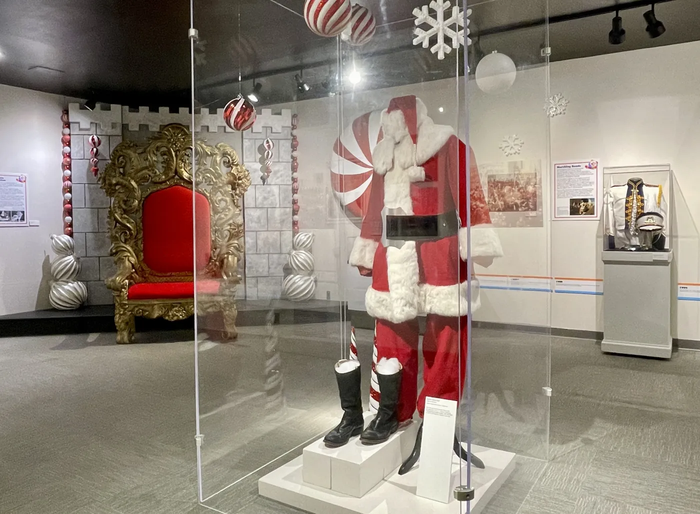Santa suit on display in a museum setting, next to a festive throne, with holiday decorations hanging above.