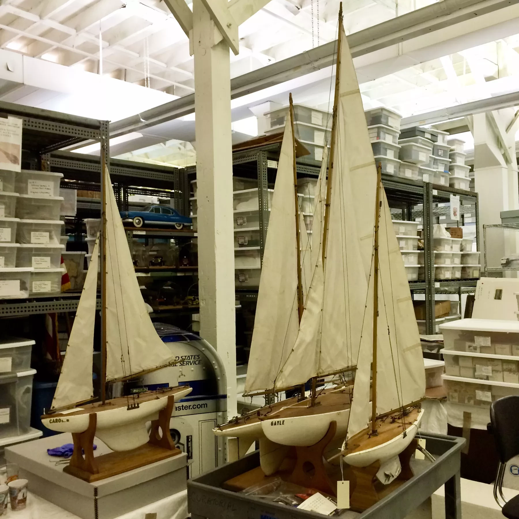 Model sailboats with white sails displayed in a storage room with shelves.