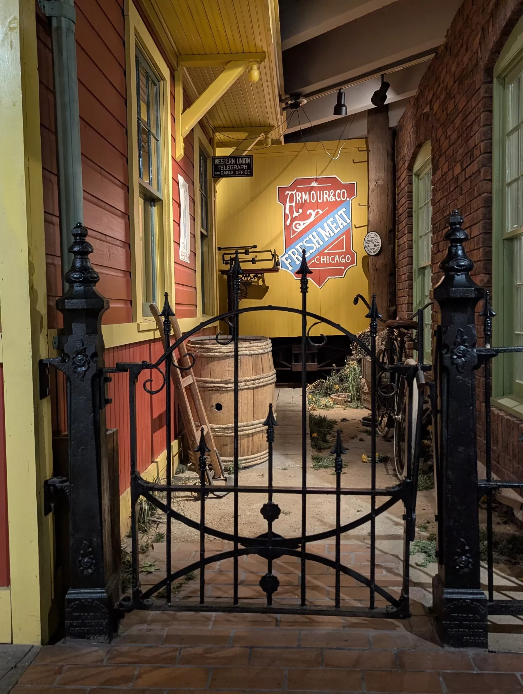 Black wrought iron gate in a vintage street scene, flanked by colorful buildings with barrels and signs behind.