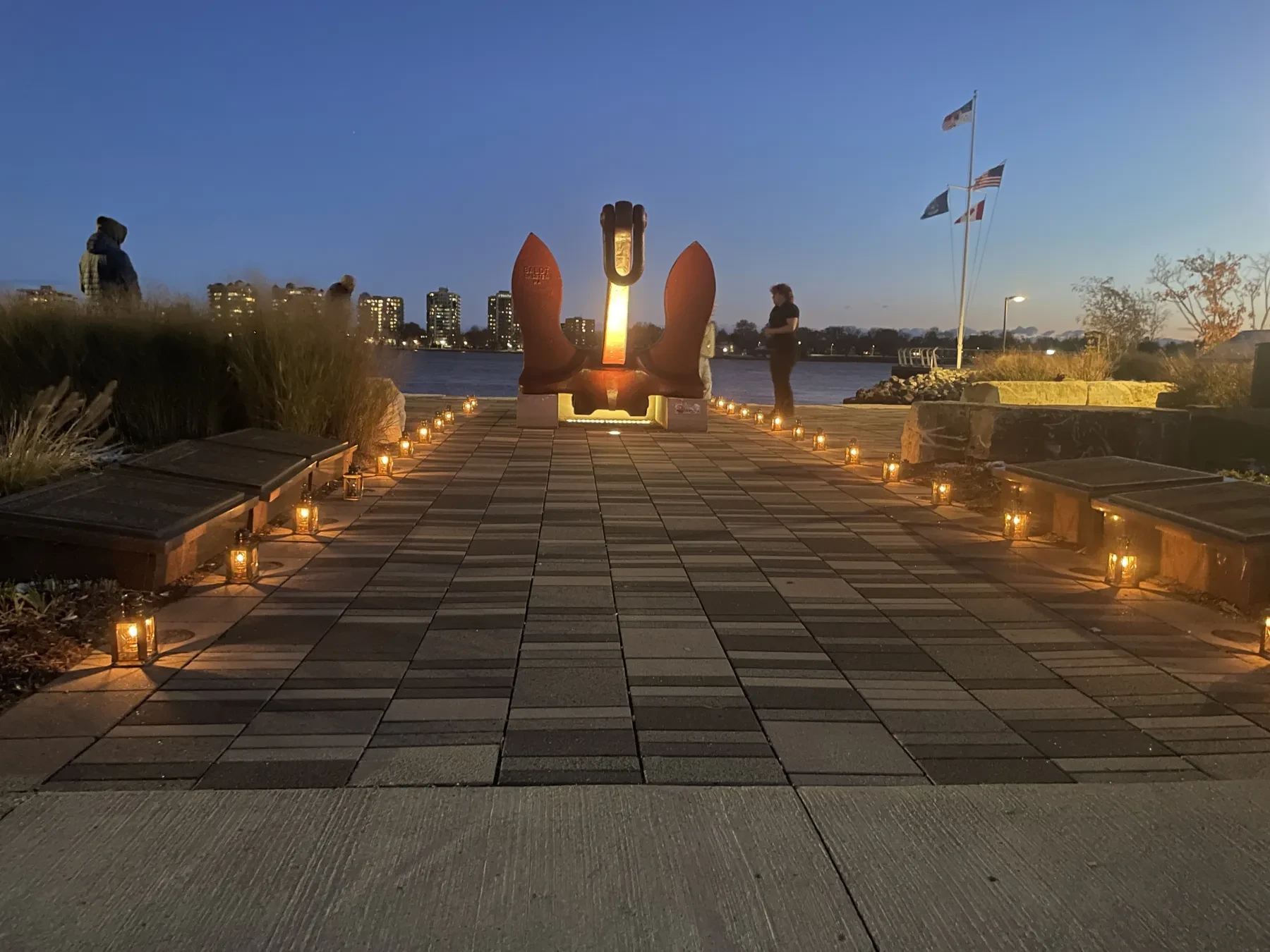 Pathway lined with candles leading to a sculpture at dusk, with flags and city skyline in the background.