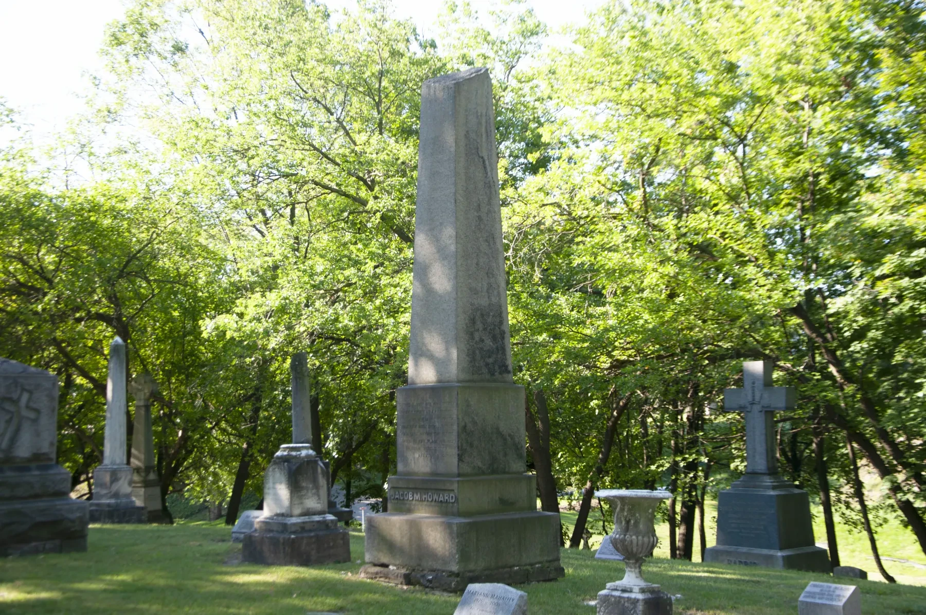 Gravestones in a cemetery under a canopy of green trees on a sunny day.
