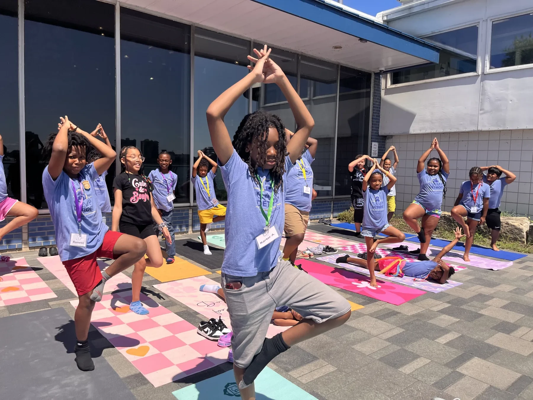 Children practicing yoga on colorful mats outside a building, holding tree pose under a sunny sky.