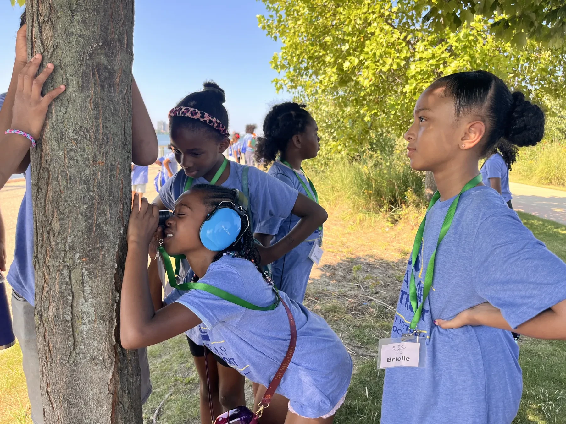 Children exploring a tree, one wearing headphones, in a sunny, grassy area.