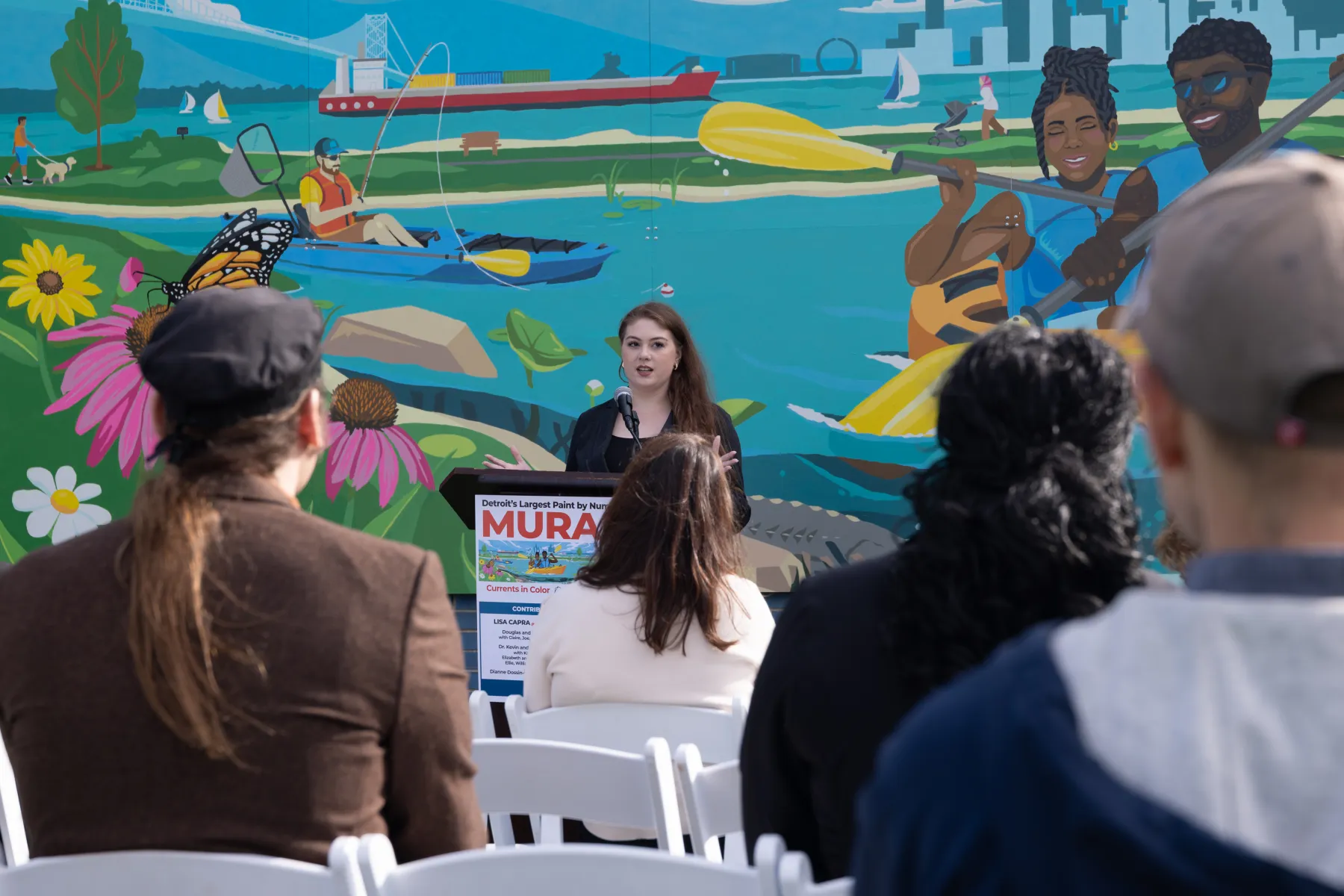A woman speaks at an outdoor event, with a colorful mural depicting kayakers and nature in the background.